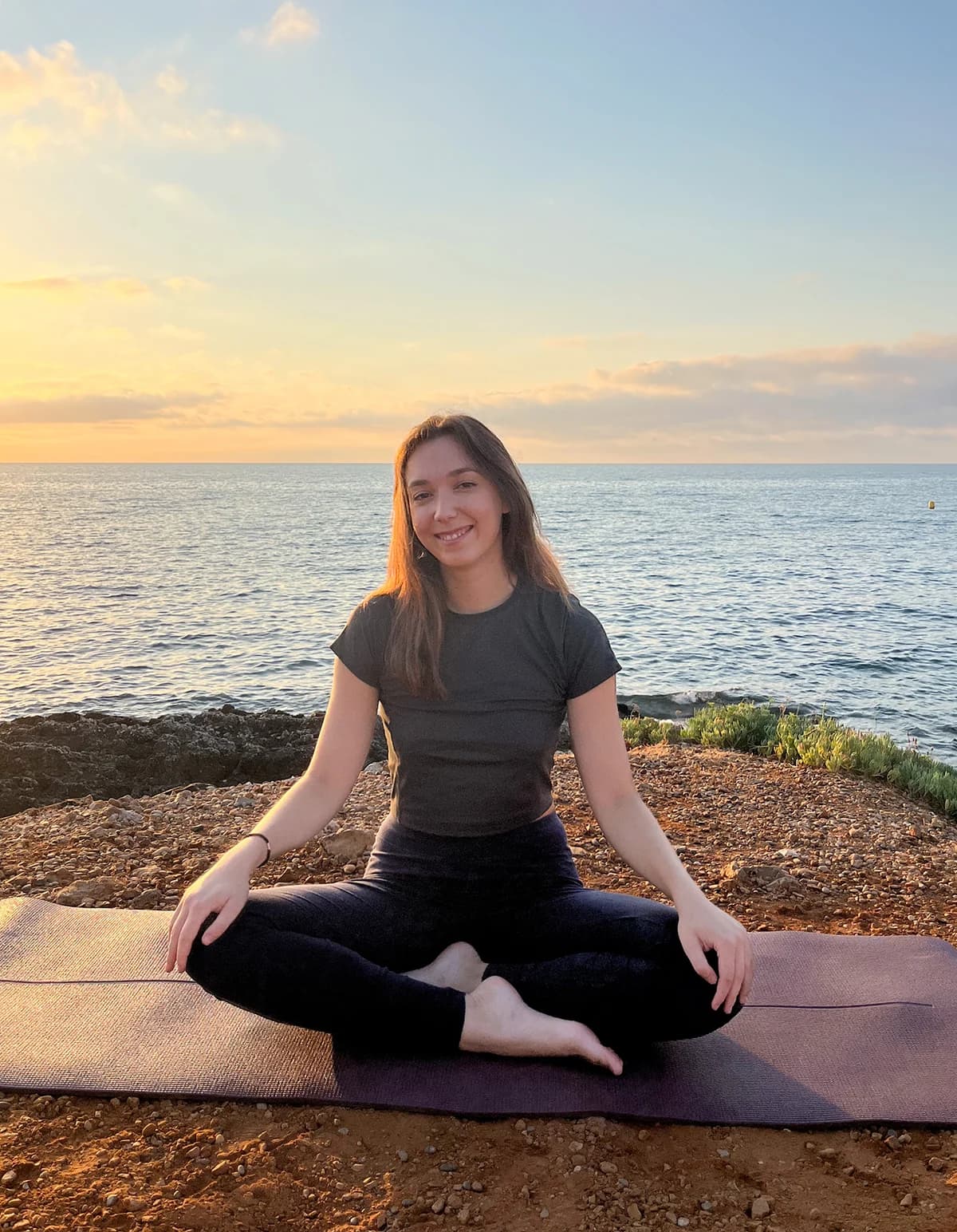 Jess souriante, assise en tailleur sur un tapis de yoga au bord de la mer au coucher du soleil